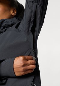 Chaqueta aislante negra con una abertura con cremallera en el lateral. Presenta una textura exterior suave y un diseño elegante y moderno.
