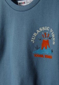 Kék pamut póló, színes grafikával, amely egy vulkánt ábrázol, a "JURASSIC VIBES" és a "COOL TIMES" felirattal fehér és narancssárga színben.