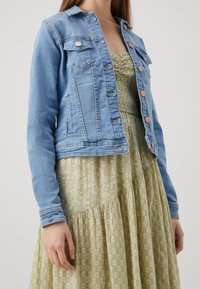Chaqueta de mezclilla de color azul claro con cierre de botones, bolsillos en el pecho y un diseño ajustado, llevada sobre un vestido verde con flores y falda de volantes.