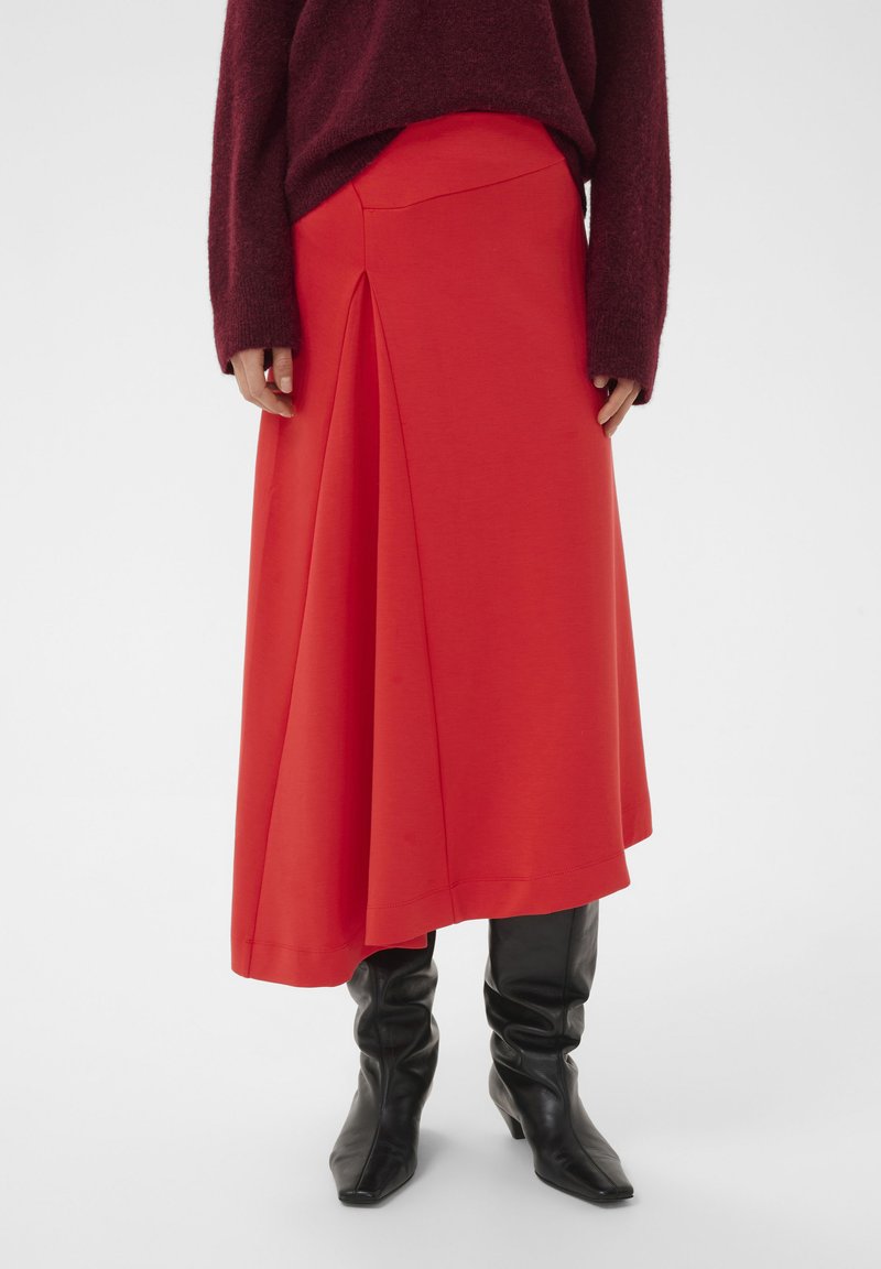 Roter Midirock mit vorderer Faltenoptik, aus glattem Stoff gefertigt. Kombiniert mit schwarzen Stiefeln mit Absatz und einem burgunderfarbenen, flauschigen Pullover.