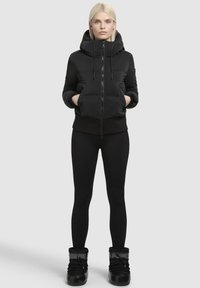 Zwarte pufferjack met capuchon, rits aan de voorkant en zijzakken, gedragen over zwarte leggings en zwarte laarzen, met een gestructureerde afwerking.
