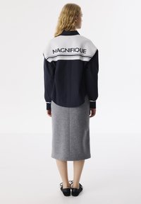 Giacca oversize blu navy con colletto bianco con la scritta "MAGNIFIQUE", abbinata a una gonna grigia fino al ginocchio e scarpe nere.