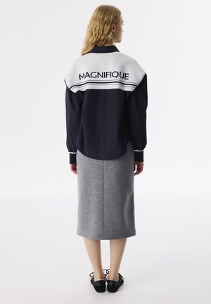 Marineblauwe oversized jas met witte kraag met de tekst "MAGNIFIQUE", gecombineerd met een grijze rok tot de knie en zwarte schoenen.