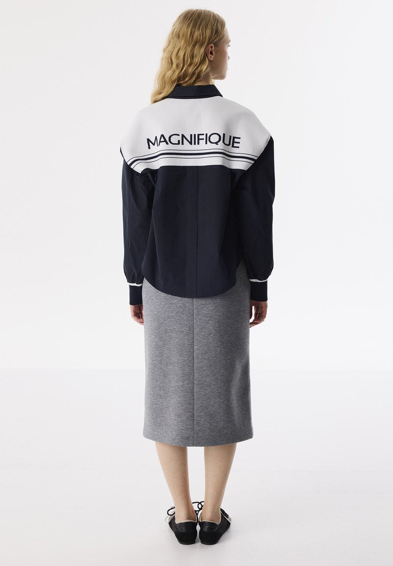 Giacca oversize blu navy con colletto bianco con la scritta "MAGNIFIQUE", abbinata a una gonna grigia fino al ginocchio e scarpe nere.