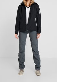 Veste softshell noire avec capuche, accompagnée d'un pantalon gris à jambes amovibles zippées. La veste possède une poche zippée et est confectionnée dans un matériau élégant et extensible.