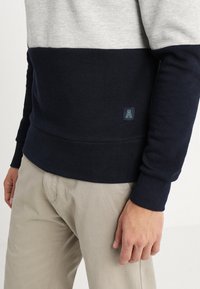 Hombre que lleva pantalones beige y un suéter con bloques de color en secciones azul marino y gris claro, con un pequeño parche de logo verde con una "A".