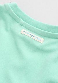 Close-up van de halslijn van een mintgroen T-shirt met een wit label dat binnenin is gestikt met de tekst "homegrown" in blauwe letters.