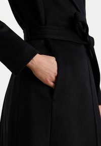 Melnais mētelis ar teksturētu audumu, apvācojošu dizainu un sasaistītu jostu, ar roku novietotu sānu kabatā redzamībai.