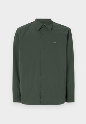Camisa de manga longa verde escura, feita de tecido suave, com gola abotoada e um logotipo discreto no lado esquerdo do peito.