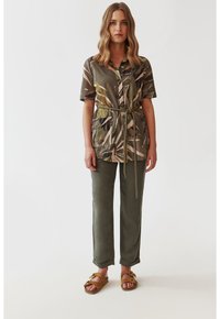 Blusa a maniche corte con stampa botanica verde, cintata in vita, abbinata a pantaloni verde oliva e sandali marroni.