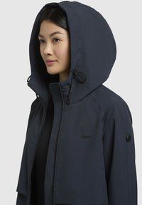 Veste à capuche bleu marine avec fermeture éclair sur le devant, col à cordon de serrage et détail du logo sur la poitrine. Tissu texturé, résistant à l'eau.