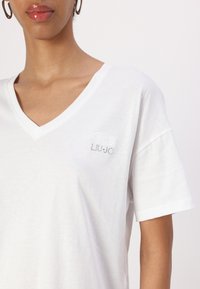 Hvid bomulds t-shirt med v-hals og korte ærmer, prydet med et diskret broderet logo med rhinsten på venstre brystområde.