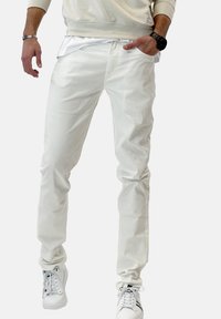 Muž v bílých slim-fit kalhotách, bílých teniskách s černými pruhy, béžovém svetru a černých hodinkách, ruka v kapse.