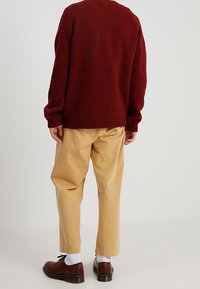 Henkilö, joka on pukeutunut marooninväriseen neuleeseen, beigepiisiin housuihin, valkoisiin sukkiin ja ruskeisiin nahkakenkiin, seisoo selin yksinkertaista taustaa vasten.