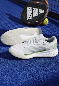 Scarpe da tennis bianche e verde chiaro su una superficie di campo blu con racchetta da padel bianca e nera e palla da tennis gialla sullo sfondo.