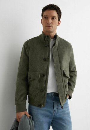 Mees, kes kannab oliivrohelise värvi nööbitud jopet valge särgi ja siniste teksade peal, hoides halli kampsunit, seisab valge seina ääres.