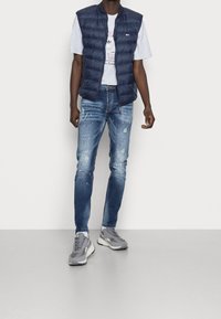 Gilet imbottito blu navy, t-shirt grigia con grafica, jeans blu strappati con spruzzi di vernice e sneakers grigie con suole bianche.