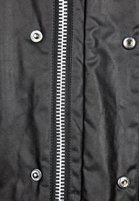 Czarna kurtka z tkaniny z widocznym srebrnym zamkiem na środku, po obu stronach ozdobiona metalowymi zatrzaskami, cechująca się gładką teksturą.