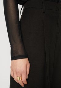 Black textured trousers with a high waist, featuring a waistband and subtle pleating, paired with a sheer black long-sleeve top. Gold ring accent.