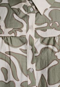 Green and beige patterned shirt featuring a button-front design, a collar, and gathered texture at the waist, with five white buttons.