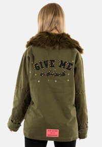 Chaqueta verde oliva con un cuello de piel sintética, acentos con tachuelas y texto bordado en la parte posterior. Incluye estrellas decorativas y un detalle de etiqueta.
