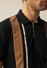 Polo noir avec un col texturé, présentant des rayures verticales marron et blanches ainsi qu'une fermeture éclair frontale. Tissu lisse avec des textures variées.