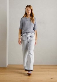 Grå stickad kortärmad tröja lager på lager över en beige skjorta med krage, bärs med ljusblå vida jeans och bruna skor på ett trägolv.