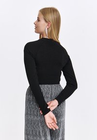 Czarny top z długim rękawem o dopasowanym kroju i prążkowanej fakturze, w zestawieniu z szarą, błyszczącą spódnicą plisowaną. Długie kolczyki stanowią dodatkowy akcent.