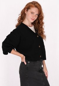 Schwarze Strickjacke mit V-Ausschnitt, drei Knöpfen und überschnittenen Schultern. Über einem hellen Oberteil getragen und mit einem hoch taillierten schwarzen Rock kombiniert.
