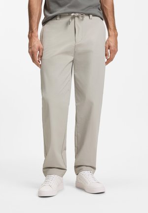 Άνδρας που φοράει ανοιχτό μπεζ παντελόνι με κορδόνια και λευκά αθλητικά παπούτσια, στέκεται με τα χέρια χαλαρά στο πλάι σε απλό φόντο.