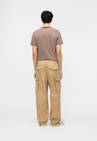 Άνδρας που φοράει μπεζ παντελόνι καμπαρντίνα και καφέ ριγέ κοντομάνικο πουκάμισο, στέκεται με το πρόσωπο προς ένα απλό λευκό φόντο.