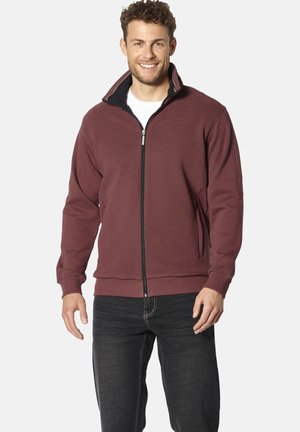 Bordeauxfarbener Zip-up-Pullover mit strukturierter Oberfläche, hohem Kragen und seitlichen Taschen. Kombiniert mit dunklen Jeans. Kontrastierender schwarzer Innenkragen.