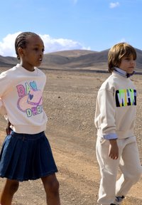 Twee kinderen lopen over een dor landschap. De een draagt een witte sweatshirt met kleurrijke tekst en een spijkerrok; de ander draagt een crèmekleurige sweatshirt en bijpassende broek.