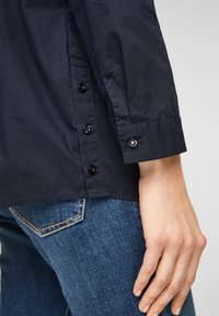 Mörkblå skjorta med knappar, gjord av en mjuk bomullstyp, med synliga knappar längs sidan och vid ärmsluten. Kombineras med blåjeans.