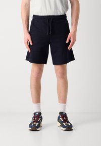 Marineblå shorts laget av strukturert stoff med elastisk midjebånd og snøring. Båret med hvite sokker og flerfargede joggesko.