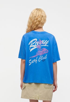 Personne blonde portant un T-shirt bleu oversize avec le texte "Roxy Surf Club" et un motif de surfeur rose dans le dos, associé à une jupe beige.