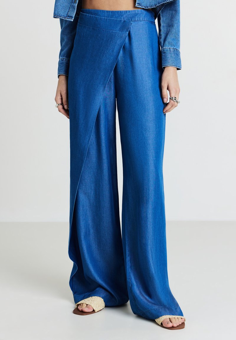 Donna che indossa pantaloni larghi blu con dettaglio incrociato frontale, giacca di jeans, anelli in argento e sandali intrecciati su uno sfondo chiaro.