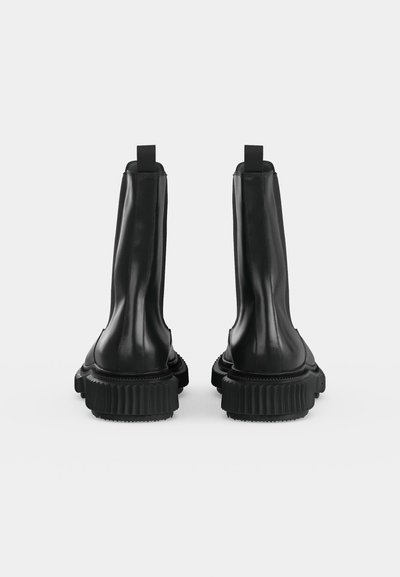 Paire de bottes hautes en cuir noir avec des semelles épaisses rainurées, vues de dos sur un fond gris clair uni.