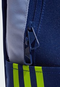 Mochila de tela azul marino con acentos grises, que presenta un doble cierre, tres rayas amarillas y una superficie texturizada. Bolsillos laterales ajustables.