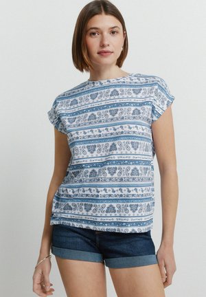 Młoda kobieta z włosami do ramion, ubrana w niebiesko-biały top z krótkim rękawem w wzór paisley oraz ciemne jeansowe, podwinięte szorty.