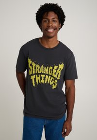 Tummanharmaa lyhythihainen t-paita, jossa kirkkaan keltainen "Stranger Things" -kuvio liekkimäisillä reunoilla. Puuvillainen materiaali, rento istuvuus.