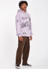 Haut à capuche lavande avec des illustrations graphiques noires ; il présente un tissu en jersey, une capuche réglable et une poche kangourou. Associé à un pantalon en velours côtelé brun.