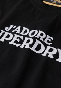 Tessuto nero con testo in rilievo bianco che legge "J'ADORE SUPERDRY" in lettere grasse, mostrando un primo piano dell'area del petto del capo.