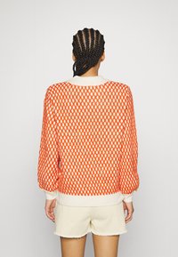 Pull orange et blanc à motifs avec une coupe décontractée, tricot texturé, poignets et ourlet côtelés, porté avec un short clair.