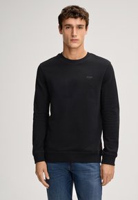 Felpa nera a maniche lunghe con collo rotondo, logo discreto sul petto e bordi a coste, abbinata a jeans blu.