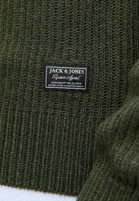 Groen gebreid trui met een geribbelde textuur en een rechthoekig zwart label met witte tekst, die het merk en details aangeeft.