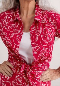 Camisa de patrón floral rojo con cintura anudada, que presenta un escote con solapa y botones. La tela tiene un acabado texturizado, superpuesta sobre una camiseta blanca.