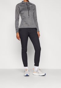 Grå långärmad topp med halvdragkedja och ficka, kombinerad med svarta smala byxor och grå sportskor med orange detaljer.