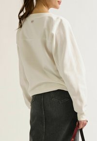 Witte sweatshirt met ronde hals en lange mouwen, met een klein label op de achterkant. Gecombineerd met een denimrok, wat een casual outfit laat zien.