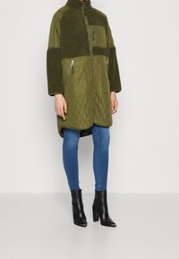 Person som bär en grön vadderad och fleecejacka med dragkedja framtill och fickor, blå tajta jeans och svarta klackboots.